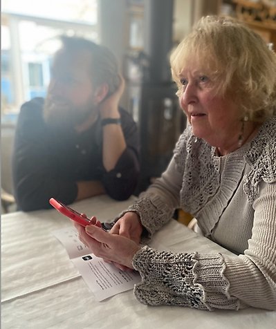 En kvinna sitter vid ett bord och använder sin mobiltelefon som fjärrkontroll. 